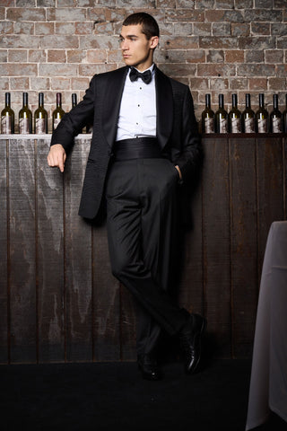 Man in a black tuxedo sitting against a brick wall with wine bottles.
