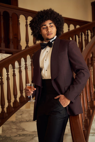 A man with a large dark afro stands on a grand staircase wearing a dark plum pinstripe tuxedo jacket, black trousers, a cream dress shirt, and a black bow tie, holding a glass of champagne.