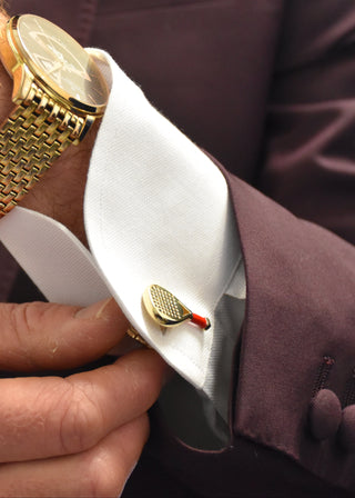 A close-up of a man's wrist and hand adjusting a gold cufflink with a small red accent on a white dress shirt cuff, wearing a gold watch and a dark plum suit jacket.