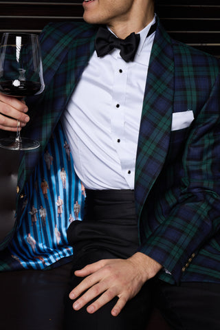 Man in formal attire with an emerald and navy plaid jacket and bow tie, holding a glass of red wine.