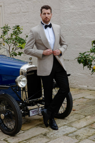 Man in a beige suit standing next to an old-fashioned car with a stone wall and plants in the background