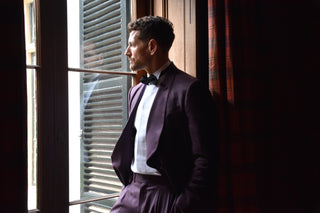 Man in a tailored plum tuxedo with a shawl lapel and black bowtie standing by an open window with blinds.