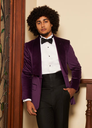A man with a large dark afro stands indoors wearing a purple velvet tuxedo jacket over a white dress shirt and black bow tie, paired with black tuxedo pants.