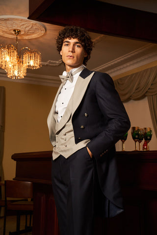 A man wearing a formal navy blue tailoring suit with a light beige waistcoat and bow tie, standing in an elegant room with a chandelier and piano.
