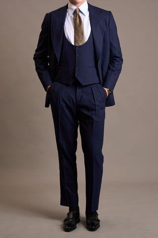 A man modeling a three-piece navy blue suit with a white dress shirt, a patterned beige tie, and black dress shoes, standing against a solid taupe studio background.