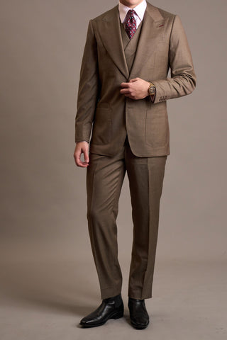 A man modeling a brown three-piece suit with a white dress shirt and a patterned purple tie, standing against a solid taupe studio background.
